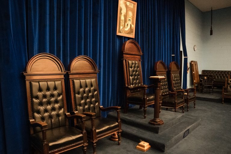 East side of a Masonic lodge room in Ottawa District 2, featuring elevated ceremonial chairs with dark leather upholstery, a wooden pedestal, and a framed historic artwork hanging against deep blue velvet curtains.