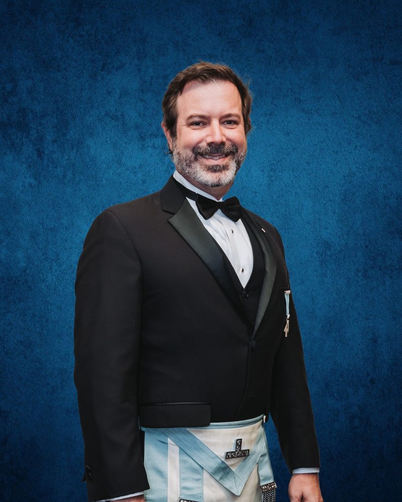 Portrait of the District Secretary of Ottawa District 2 wearing formal Masonic attire with light-blue and white regalia, standing against a textured blue background, representing administrative leadership within the Freemasons of Ontario.