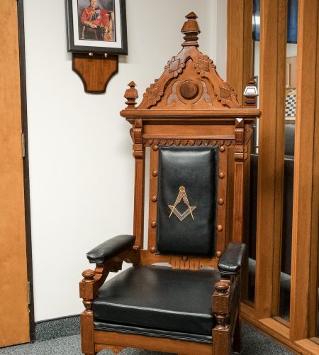 Intricately carved wooden Masonic chair with black leather upholstery and the square-and-compasses emblem, set within an Ottawa District 2 lodge anteroom, with a framed portrait of a distinguished Mason hanging above.